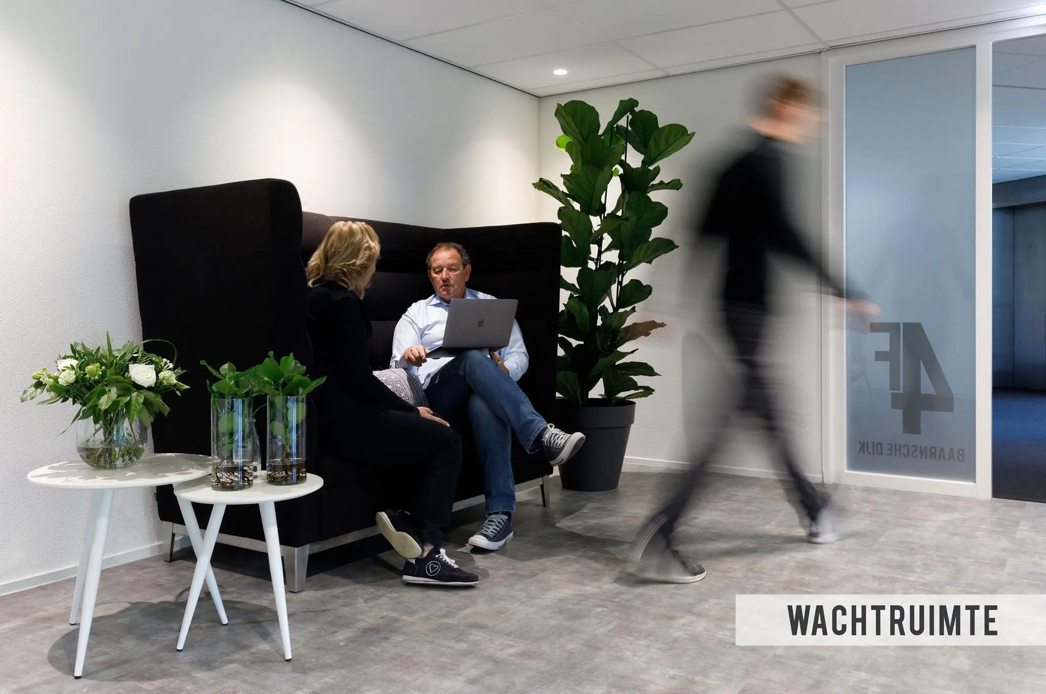 Twee mensen zitten in een wachtruimte aan de Baarnsche Dijk, met een laptop op schoot en een voorbijlopende persoon op de achtergrond.